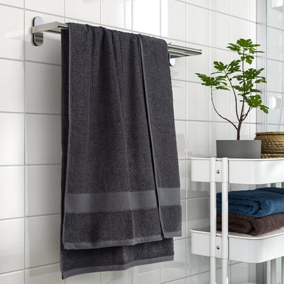 Dark grey VITBACKEN bath towel hanging on silver rail, larger than folded towels on white shelf with plant.