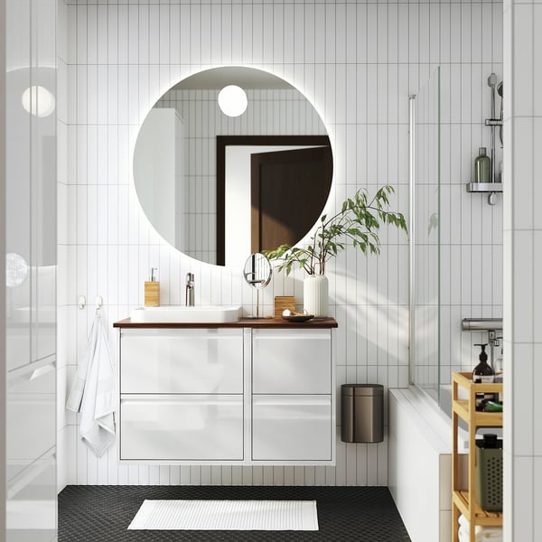 Moderno bagno bianco con specchio rotondo, elegante lavabo e doccia in vetro. Asciugamani bianchi e piante decorano.