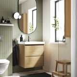 Bagno moderno con lavabo ÄNGSJÖN, due cassetti, piastrelle verde salvia, lavandino nero, specchio rotondo, pianta.