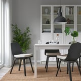 Sala da pranzo moderna con pareti bianche, sedie grigie, tavolo bianco, lampada nera, piante e libri.