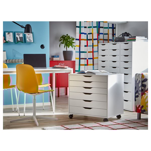 Ufficio moderno con contenitori ALEX bianchi su ruote, sedia gialla, armadio rosso, parete blu e tappeto colorato.