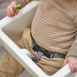 Bambino in seggiolone ANTILOP, indossa una camicia a righe, cuori sui pantaloni, cucchiaio verde.