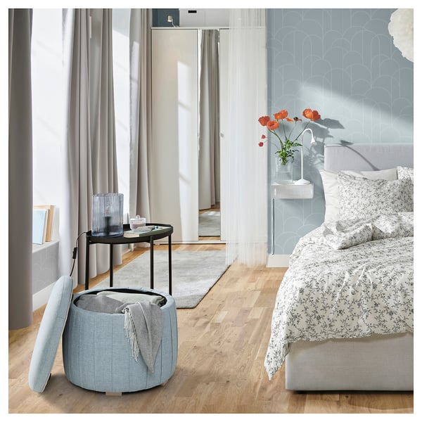 Camera da letto a motivi blu con pavimento in legno e grande finestra. Letto floreale, pouf rotondo, tavolino con papaveri e candela.
