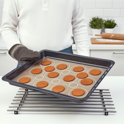 Persona che tiene la teglia con biscotti circolari, posta su una gratella di raffreddamento. Tappetino BAKTRADITION visibile, che favorisce una cottura uniforme e un facile rilascio.