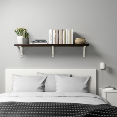 Camera da letto moderna con mensola BERGSHULT a parete, che espone libri, piante e un cesto intrecciato sopra un letto bianco con cuscini grigi.