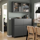 Cucina moderna con mobili contenitori BESTÅ grigio bicolore, sedie in vimini e vetrine con piatti e tazze.
