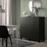 Spazio abitativo moderno con mobile BESTÅ nero, parete verde, scrivania bianca, specchio, lampade e piante.