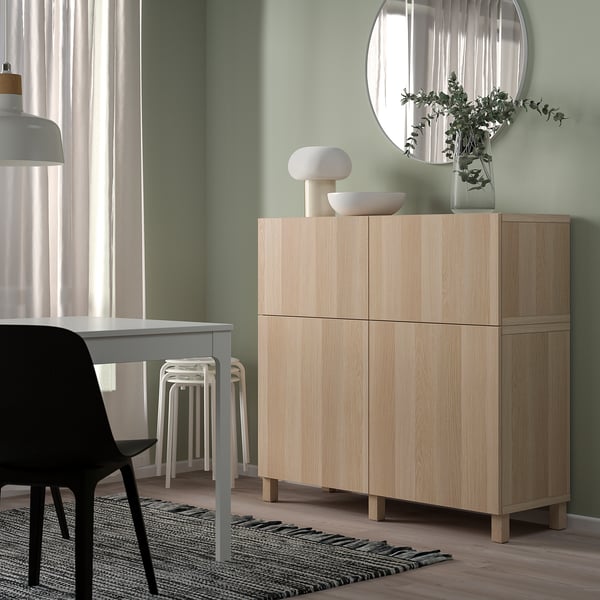Armadio BESTÅ in legno chiaro contro parete verde, con specchio rotondo, sedia nera e sgabelli bianchi.