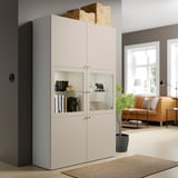 Mobile BESTÅ beige con ante in vetro e cestello. Vicino alla finestra, alla pianta e alla sedia.