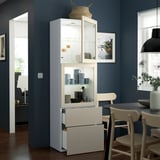 Una moderna sala da pranzo con un mobile BESTÅ bianco con ante e cassetti in vetro, con tazze e decorazioni, addossato a pareti blu scuro.