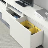 Porta tv BESTÅ bianco con cassetto aperto con cestino giallo e telecomando, oltre a un ripiano chiuso con libri e TV a schermo piatto sulla parte superiore.