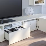 Mobile tv BESTÅ bianco con cassetto aperto per i controller di gioco e le console, ripiano chiuso con cavi a vista e lampade sulla parte superiore.