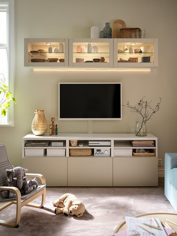 Soggiorno moderno con mobile tv BESTÅ bianco, tv a schermo piatto e mensole integrate che espongono vasi e decorazioni. Pareti e moquette beige chiaro.