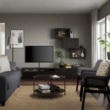 Soggiorno moderno con porta tv BESTÅ, tv a schermo piatto montata, divano e poltrona grigi, pensili in legno scuro, tavolino rotondo e decorazioni.