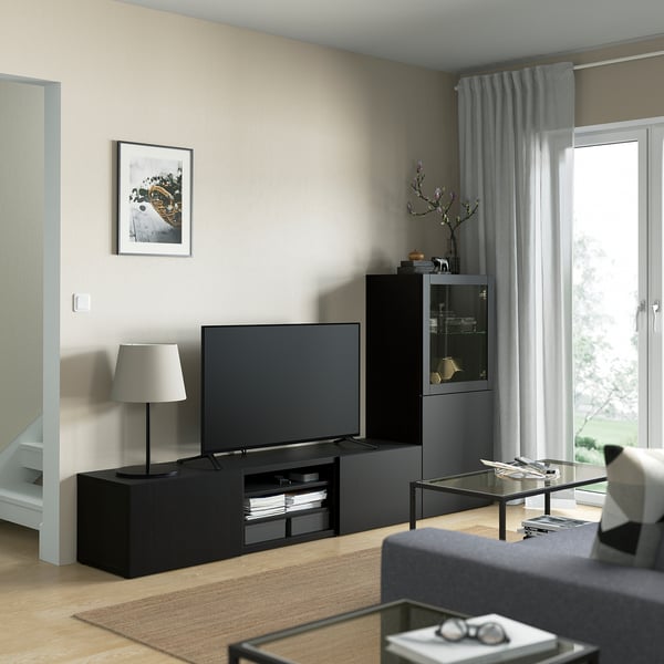 Soggiorno moderno con porta tv BESTÅ, grande tv, tavolino in vetro e tende grigie.