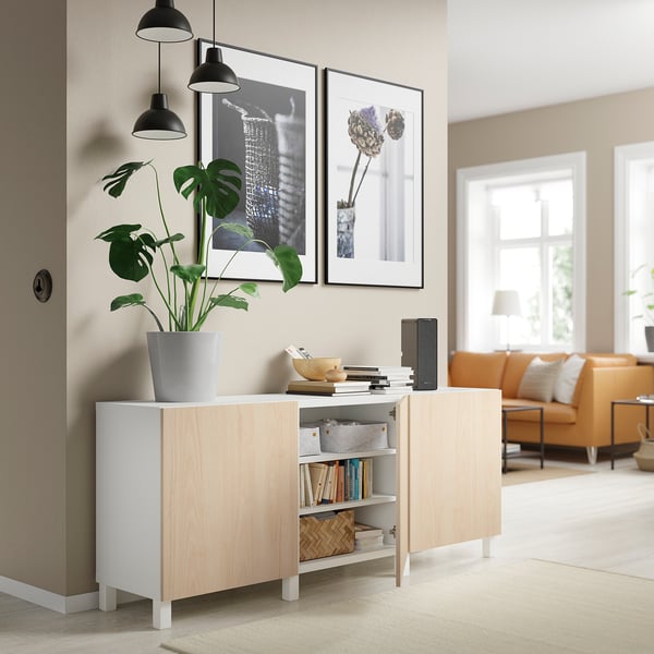 Soggiorno moderno con credenza BESTÅ bianca e in legno chiaro, pianta d'appartamento, lampade a sospensione, opere d'arte incorniciate e divano arancione.