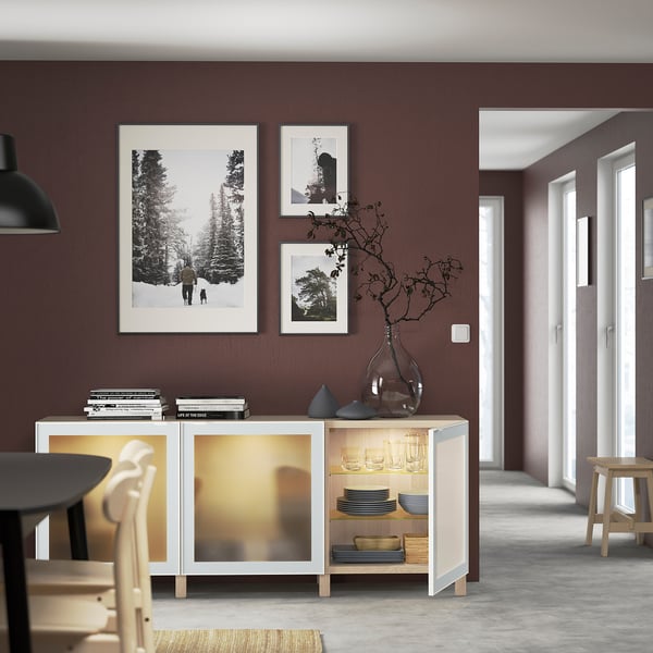 Moderna sala da pranzo con credenza BESTÅ, che conserva piatti, libri e un vaso con rami, sotto foto della natura in bianco e nero su una parete marrone scuro.