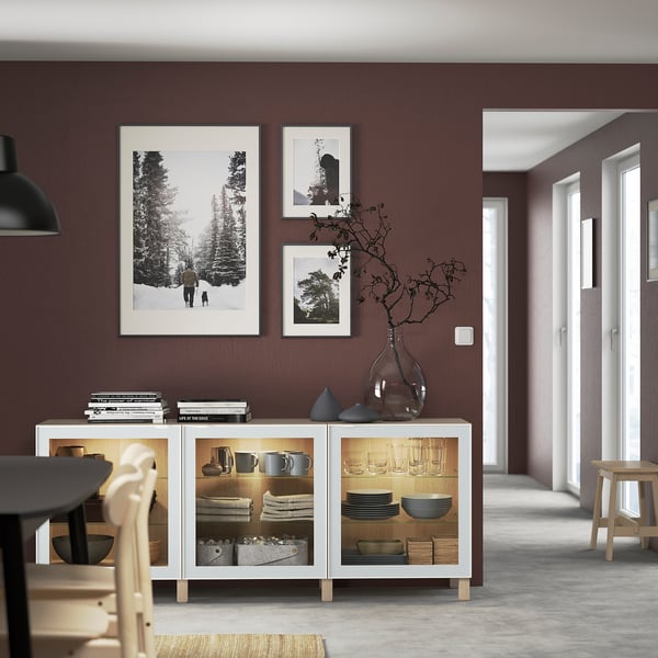 Credenza bianca BESTÅ con ante in vetro, che espone piatti e vasi, sotto tre opere d'arte incorniciate su una parete color moka.