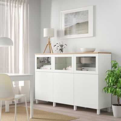 Armadio in vetro bianco BESTÅ con lampada, ciotola, pianta, tra finestra e tavolo da pranzo.