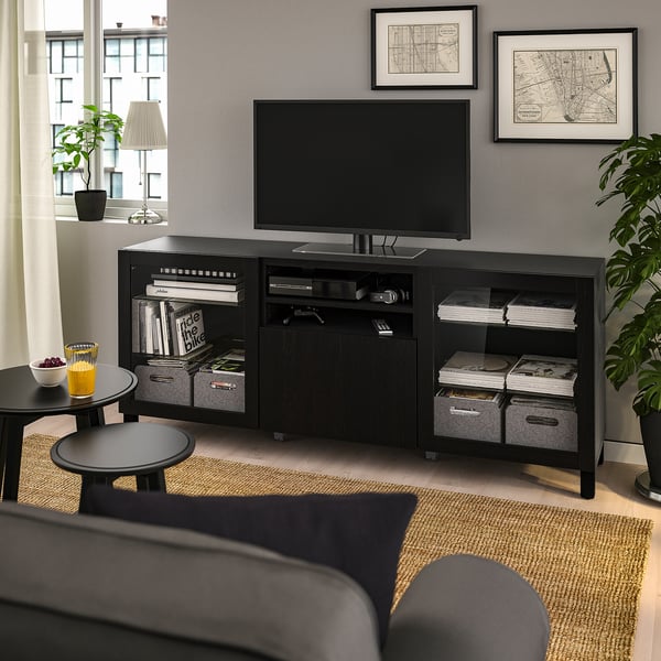 Soggiorno moderno: console tv BESTÅ, schermo piatto, divano grigio, tavolini con bevande e frutta, grande finestra, piante, mappe, libri, scatole su console.
