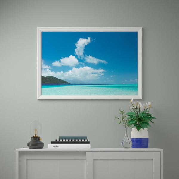 Foto incorniciata della spiaggia tropicale, consolle bianca con lampada, libri e vaso floreale.