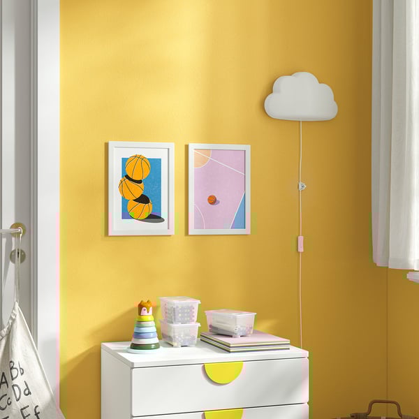 Muro giallo con opere d'arte bianche, palloni da basket, forme pastello. Comò bianco, giocattoli, libri, lampada nuvola.