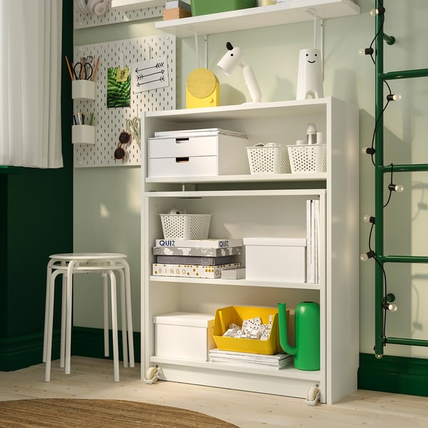 Libreria BILLY bianca con scatole e giochi, annaffiatoio verde, sgabello bianco e mensola a muro con oggetti accanto all'anta dipinta di verde.