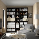 Libreria BILLY nera in camera rivestita in legno con libri e contenitore. Divano grigio, lampada, foto e puzzle nelle vicinanze.