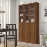 Grande libreria BILLY in legno marrone con ante in vetro, che contiene piatti e bicchieri, accanto a sedie nere e tavolo da pranzo, parete bianca e fondale per tende.