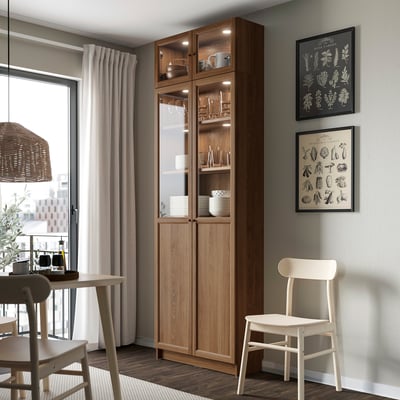 Un moderno angolo pranzo, un'alta libreria BILLY in legno piena di piatti e bicchieri, affiancata da stampe botaniche e una sedia bianca.