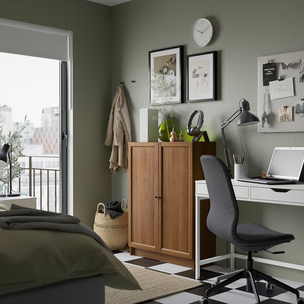 Accogliente camera da letto con libreria BILLY, scrivania, sedia, lenzuola verde oliva e parete verde con opere d'arte e orologio.