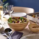 Grande ciotola di legno di insalata sul tavolo. Vicino a bicchieri da vino, piatti e ciotola più piccola con semi. Accompagnato da utensili da portata e tovaglioli.