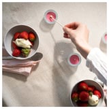 La mano accende una candela profumata alla fragola vicino a una ciotola di fragole e gelato. Superficie in legno chiaro, coppe per candele rosa.