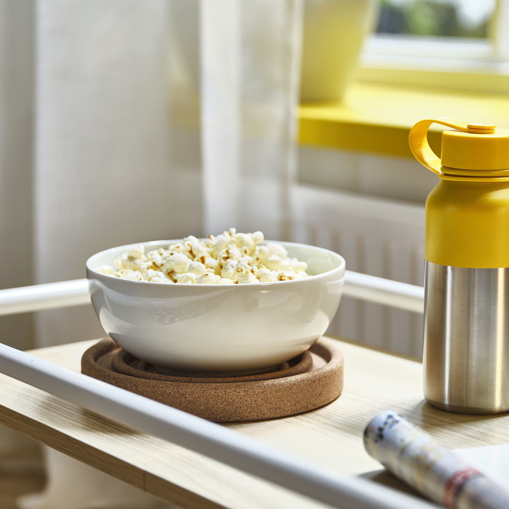 Il sottobicchiere in sughero BRÄNNBOLL contiene una ciotola bianca di popcorn accanto a una fiaschetta gialla. Il design circolare si adatta perfettamente alla ciotola, evitando fuoriuscite.