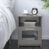 Moderno comodino grigio BRIMNES con ripiani aperti e cassetto vicino a un letto bianco. Contiene una pila di libri, una tazza di caffè bianca e una sveglia nera.