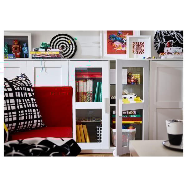 Soggiorno moderno con armadio bianco BRIMNES, sedia rossa, cuscini in bianco e nero, vari oggetti decorativi sugli scaffali e una tazza di caffè su un tavolo.