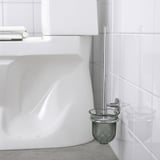 Un portascopino cromato montato su una parete del bagno piastrellata bianca, che nasconde i fissaggi e mantiene lo spazzolino a portata di mano e sollevato dal pavimento.