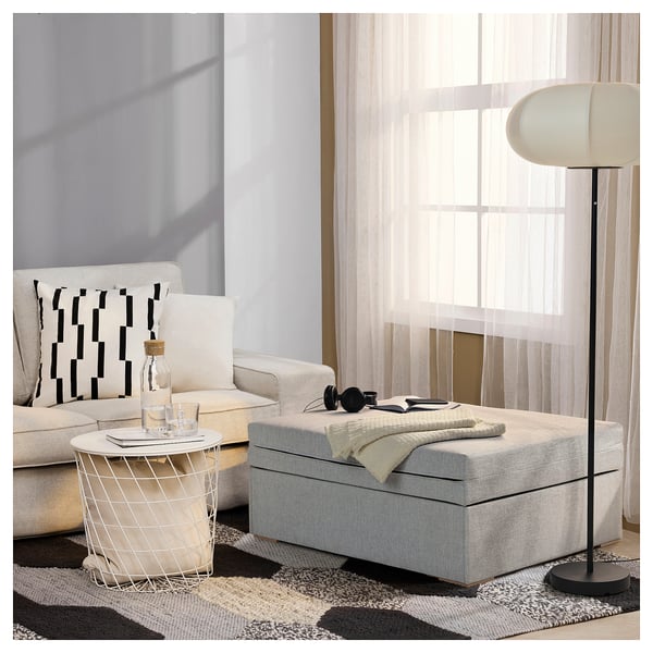 Soggiorno moderno con divano bianco, cuscino a motivi geometrici, tavolino in filo metallico, pouf portaoggetti in tessuto grigio e lampada da terra.