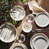 DAGGASTER piatti, ciotole e bicchieri in gres beige chiaro. Finitura rustica e maculata. Piatti più grandi circondano ciotole più piccole su un tavolo di legno.