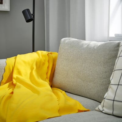 Giallo brillante DAGGKÅPOR fleece plaid su divano grigio, morbido, rettangolare, accanto a grande cuscino bianco.