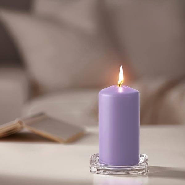 Una candela DAGLIGEN color lavanda accesa su una base di vetro trasparente si trova su un tavolo, più piccola dei libri vicini.
