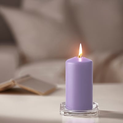 Una candela DAGLIGEN color lavanda accesa su una base di vetro trasparente si trova su un tavolo, più piccola dei libri vicini.