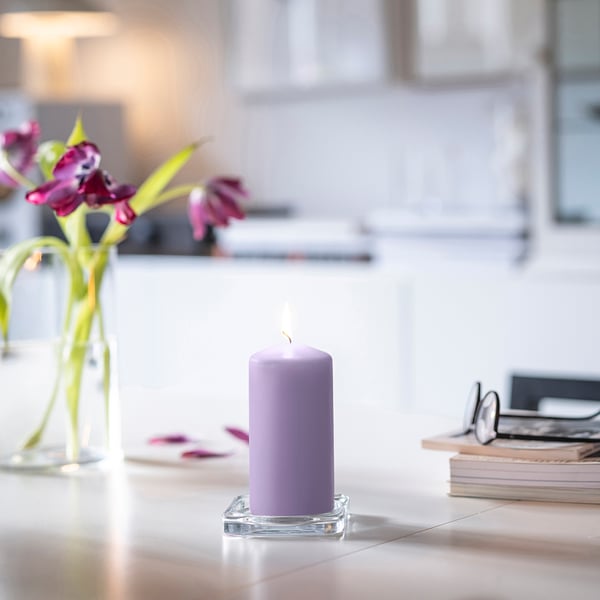 Una piccola candela DAGLIGEN color lavanda accesa si trova su un portabicchieri, accanto a un vaso con fiori e libri impilati su un tavolo bianco.