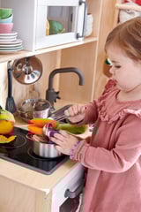 Una bambina gioca con la cucina giocattolo DUKTIG, tagliando verdure arancioni su un fornello nero, indossando un maglione rosa.