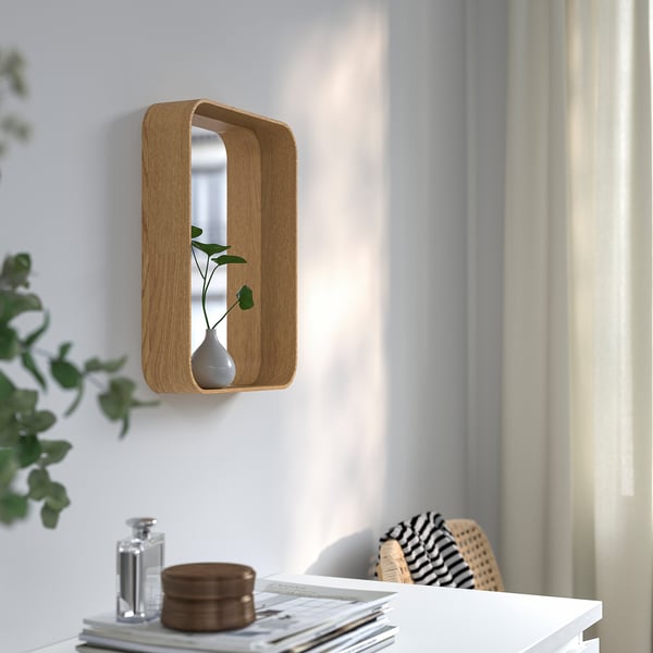 Uno specchio incorniciato in legno con mensola contiene un vaso, montato su una parete bianca.