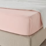Un letto ben fatto con un lenzuolo rosa chiaro, leggermente stropicciato, su un materasso beige. Il lenzuolo è liscio e sembra essere di cotone.