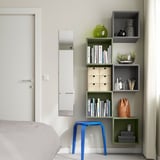 Camera da letto moderna con contenitore EKET in grigio e verde, che contiene libri e decorazioni, con uno sgabello blu accanto al letto.
