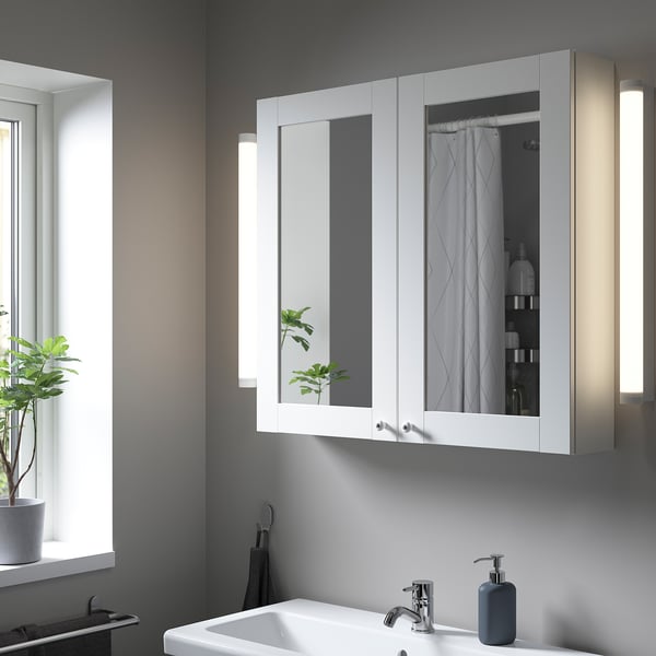 Bagno moderno con pensile bianco ENHET. Le grandi ante a specchio valorizzano lo spazio; case articoli da toeletta. Parete grigia e lavandino sottostante.