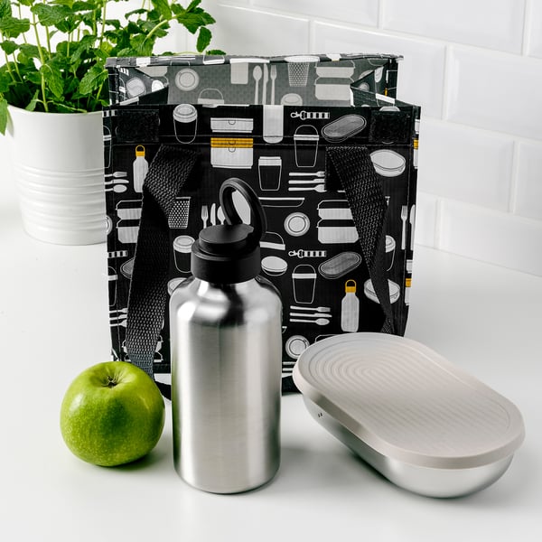 Borsa fantasia nera con utensili da cucina, bottiglia d'acqua d'argento, mela verde e contenitore in metallo aperto su bancone bianco.