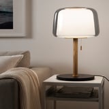Moderna lampada EVEDAL sul tavolo, paralume in vetro e supporto in legno, vicino a un accogliente divano grigio, design elegante ed equilibrato, si adatta bene allo spazio abitativo.